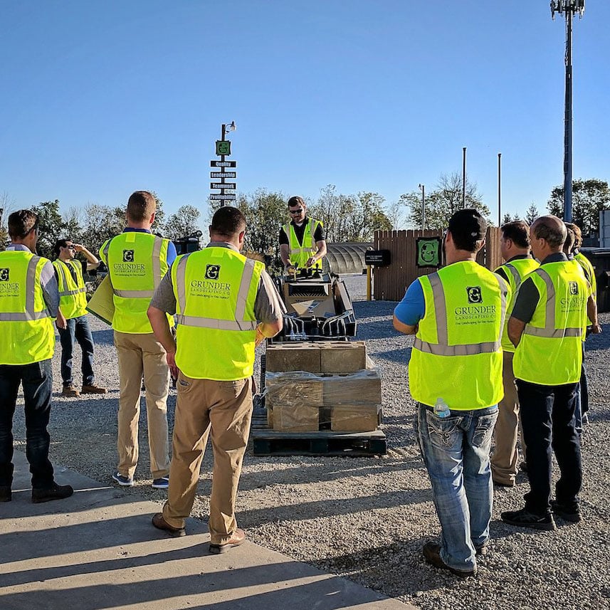 Landscaping crew training on landscape equipment. One employee on a skid steer moving large hardscape material.